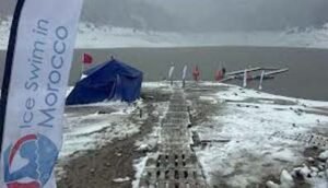 Snowy landscape with a wooden path leading to a lake, a blue tent, flags, and a banner promoting ice swimming in Morocco.