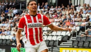PSV Eindhoven player in a red and white striped kit celebrates on the pitch, with fans and stadium seats visible in the background.