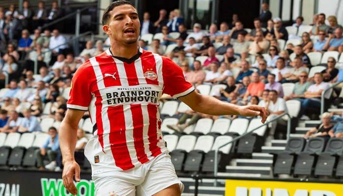 PSV Eindhoven player in a red and white striped kit celebrates on the pitch, with fans and stadium seats visible in the background.