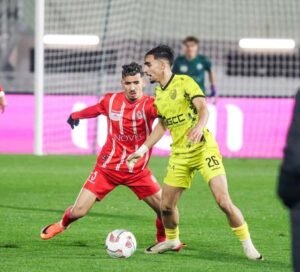 Een in rood-wit geklede speler in actie tegen een in geel geklede tegenstander tijdens een competitieve voetbalwedstrijd, met een wazig publiek op de achtergrond.