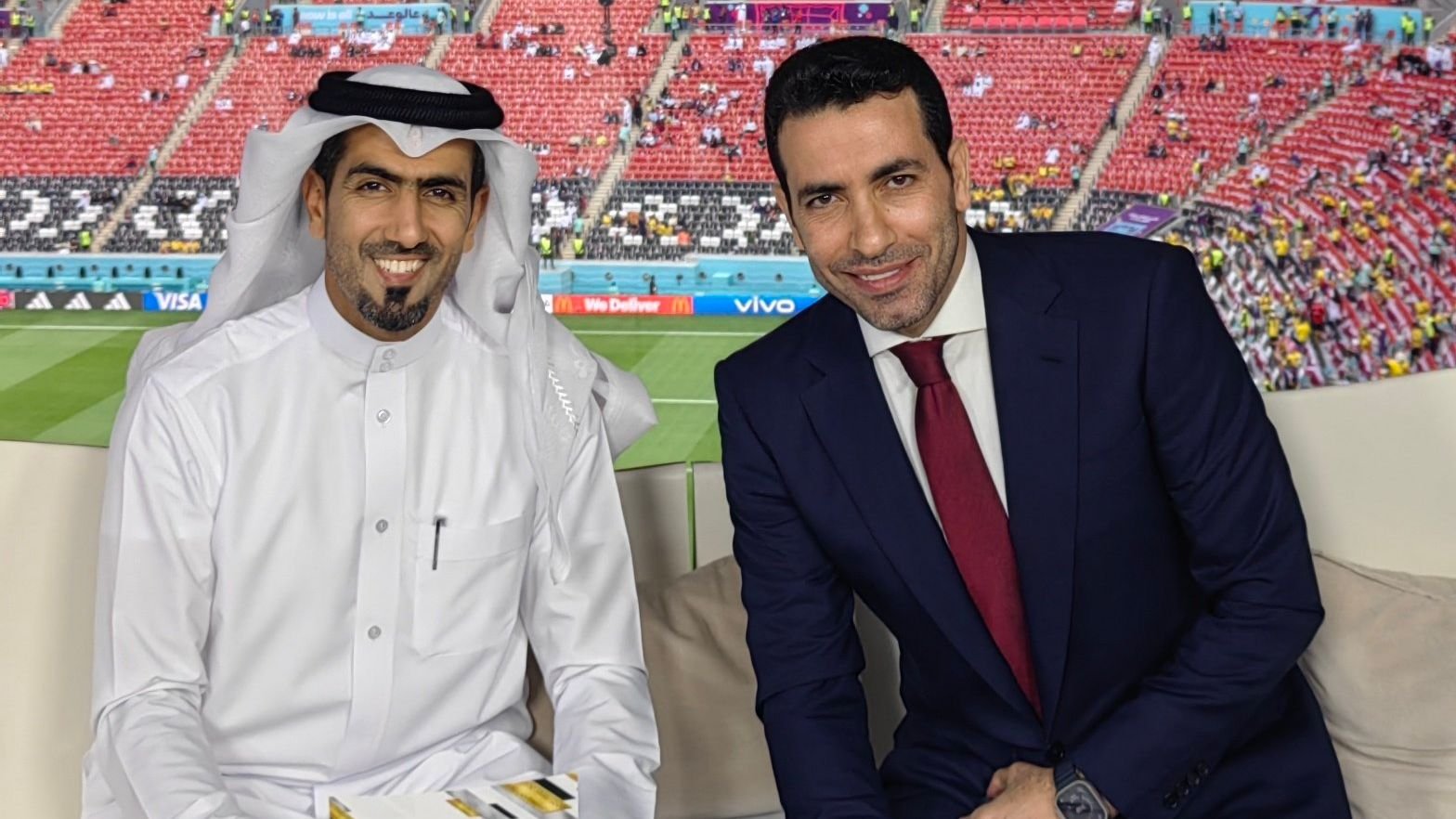 مذيعان في إطار ملعبي، أحدهما يرتدي الزي الأبيض التقليدي والآخر يرتدي بدلة كحلية، يبتسمان على خلفية كرة قدم.