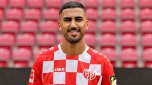 لاعب كرة قدم مغربي يبتسم بثقة، مرتدياً قميصاً مربعات أحمر وأبيض، يقف أمام مقاعد ملعب فارغة.