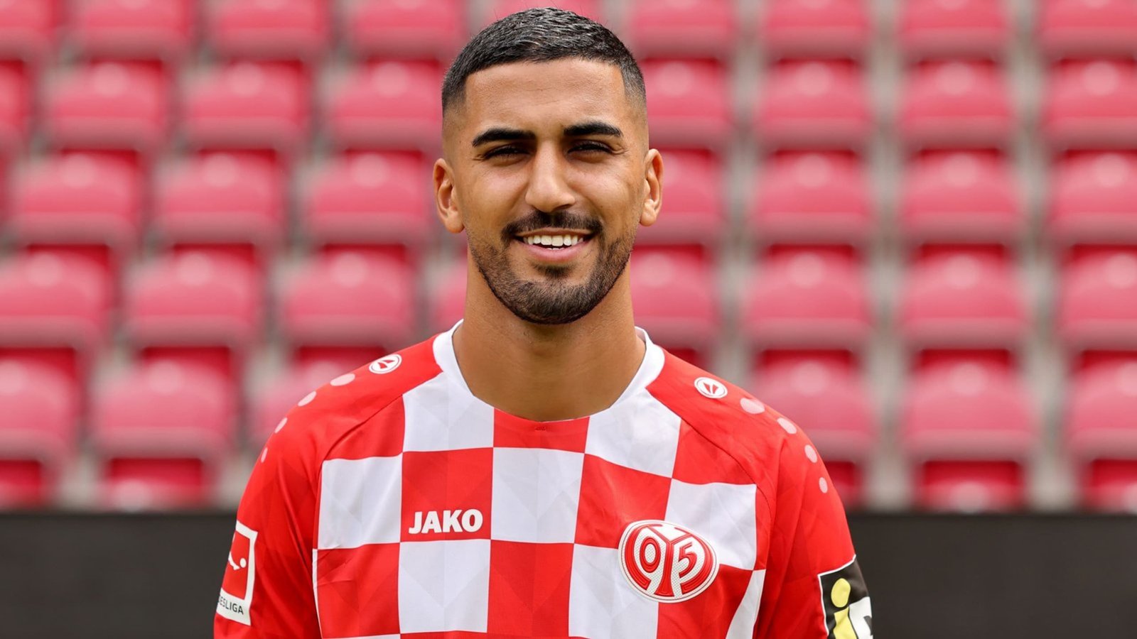 لاعب كرة قدم مغربي يبتسم بثقة، مرتدياً قميصاً مربعات أحمر وأبيض، يقف أمام مقاعد ملعب فارغة.