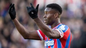 Crystal Palace player in a red and blue striped kit, gesturing passionately during a match, with gloves on, against a blurred crowd background.