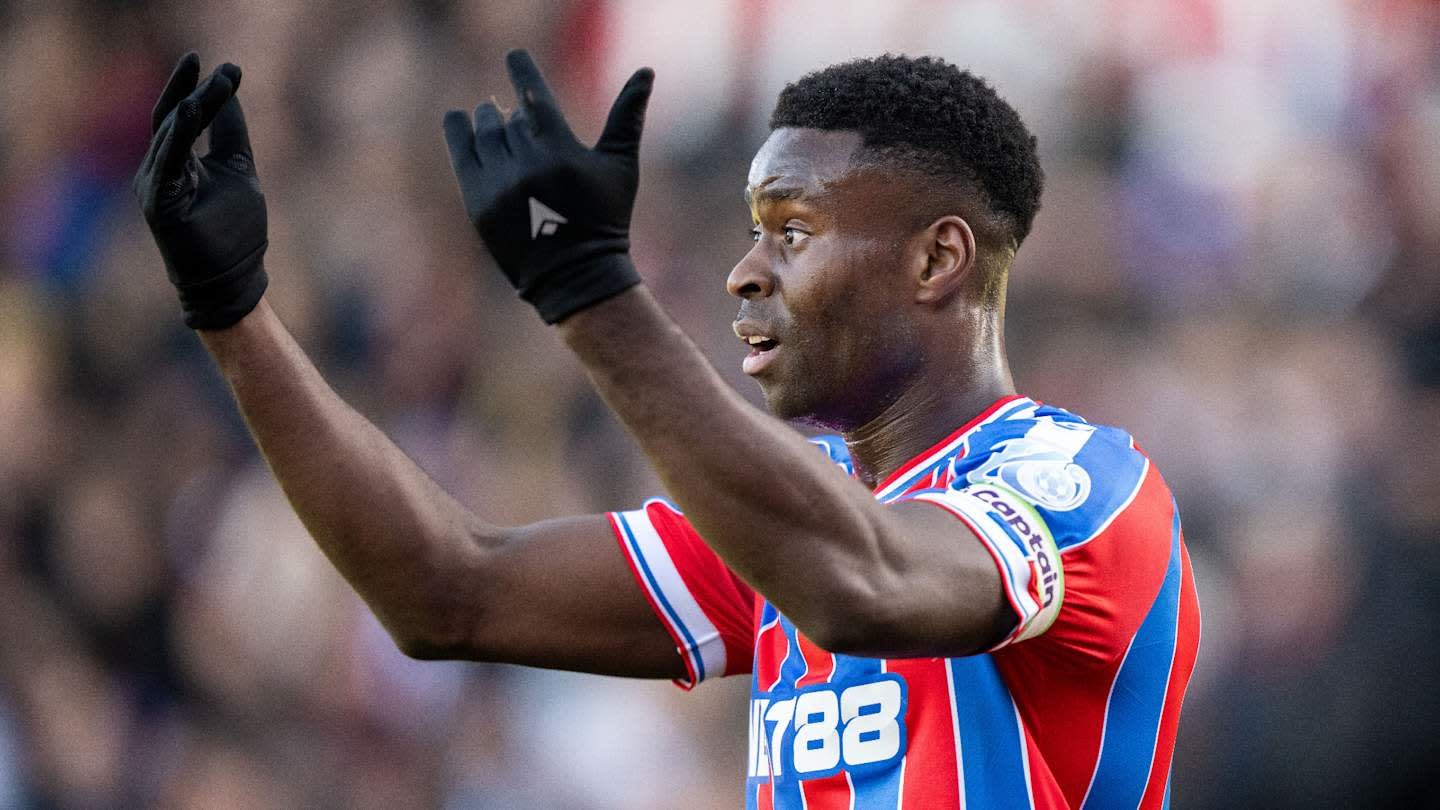 Crystal Palace player in a red and blue striped kit, gesturing passionately during a match, with gloves on, against a blurred crowd background.