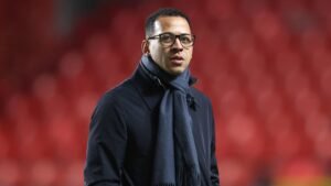 Homme portant un manteau sombre et une écharpe, avec des lunettes, debout devant un fond rouge flou, dégageant une humeur contemplative liée au football.