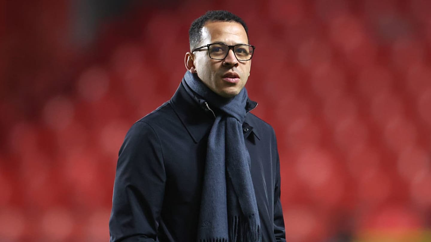 Homme portant un manteau sombre et une écharpe, avec des lunettes, debout devant un fond rouge flou, dégageant une humeur contemplative liée au football.