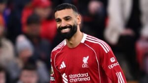 Mohamed Salah sourit en portant le maillot rouge de Liverpool, affichant la fierté de l'équipe contre un arrière-plan flou de fans.