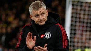 Ole Gunnar Solskjaer, gekleed in een Manchester United-trainingspak, gebaart met een bezorgde uitdrukking tijdens een wedstrijd. Op de achtergrond is een vervaagd voetbaldoel te zien.