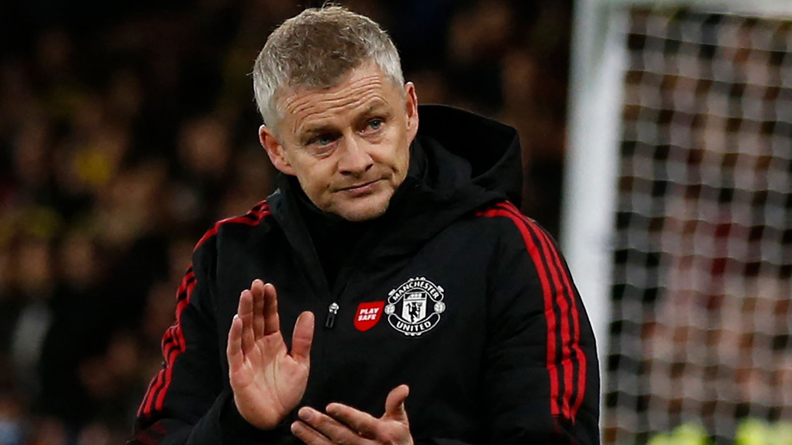 Ole Gunnar Solskjaer, gekleed in een Manchester United-trainingspak, gebaart met een bezorgde uitdrukking tijdens een wedstrijd. Op de achtergrond is een vervaagd voetbaldoel te zien.