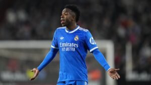 Vinícius Jr. in een levendig blauw Real Madrid-tenue, toont frustratie tijdens een wedstrijd, met het stadion wazig op de achtergrond.