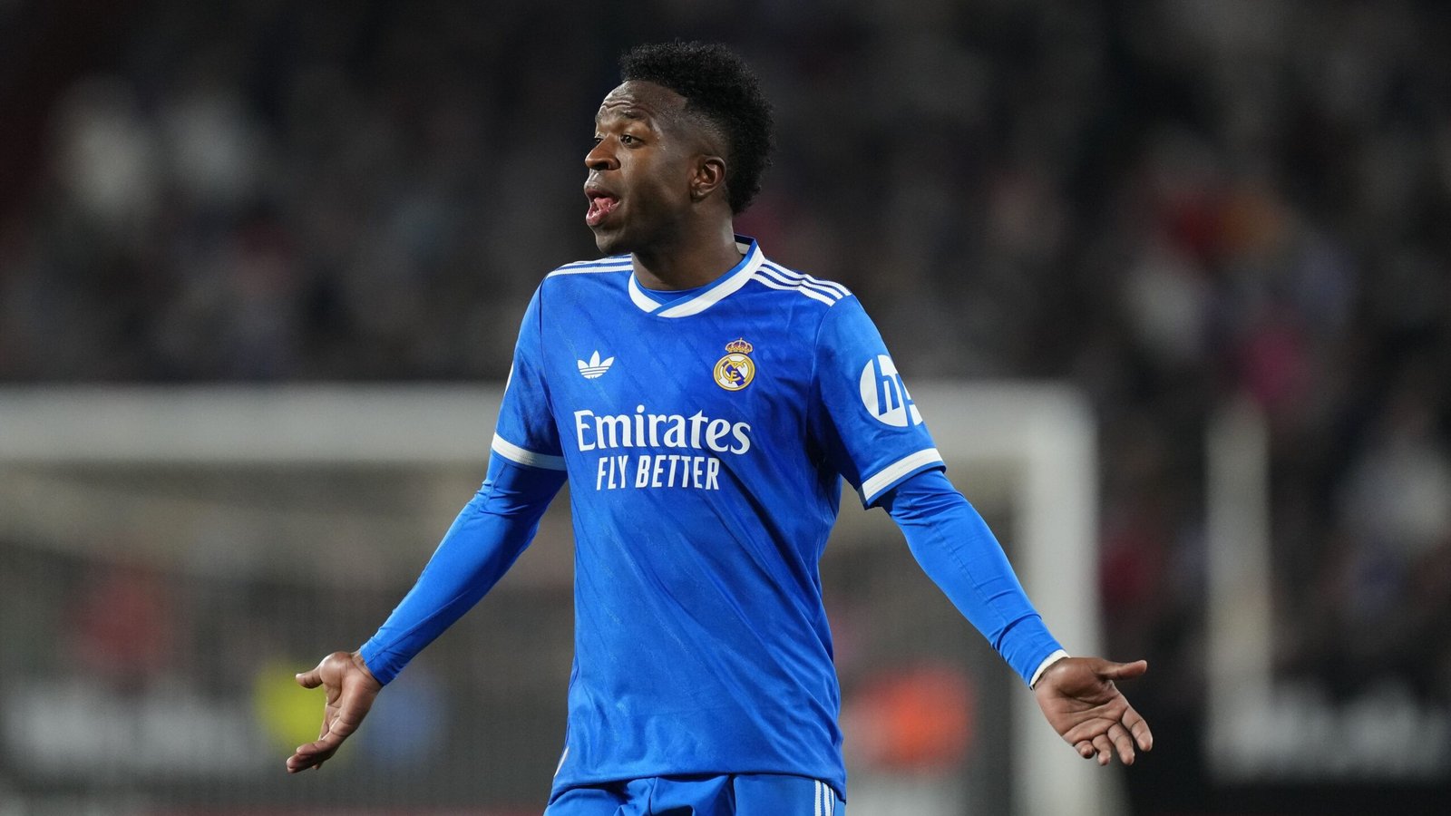 Vinícius Jr. in een levendig blauw Real Madrid-tenue, toont frustratie tijdens een wedstrijd, met het stadion wazig op de achtergrond.