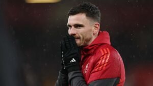 Andy Robertson, wearing a red Liverpool training jacket, reacts with his hands to his face, rain visible in the background.