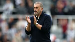 Ange Postecoglou, in een marineblauwe trui, applaudisseert vol overgave tijdens een wedstrijd, met vervagende fans en stadionachtergrond.