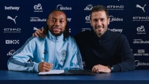 Antoine Semenyo sourit aux côtés d’un représentant du club lors de la signature de son contrat avec Manchester City, vêtu d’une veste d’entraînement bleu clair.
