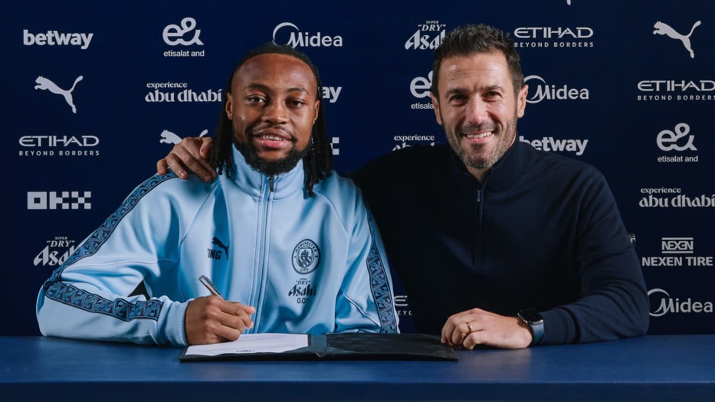 Antoine Semenyo sourit aux côtés d’un représentant du club lors de la signature de son contrat avec Manchester City, vêtu d’une veste d’entraînement bleu clair.