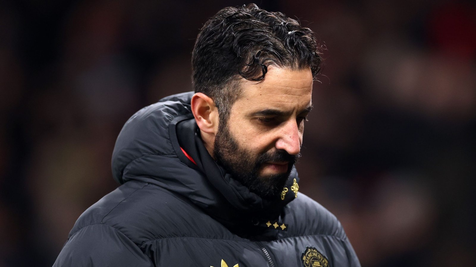 Ruben Amorim apparaît pensif, portant une veste matelassée noire, avec un stade flou en arrière-plan, reflétant une atmosphère de match tendue.