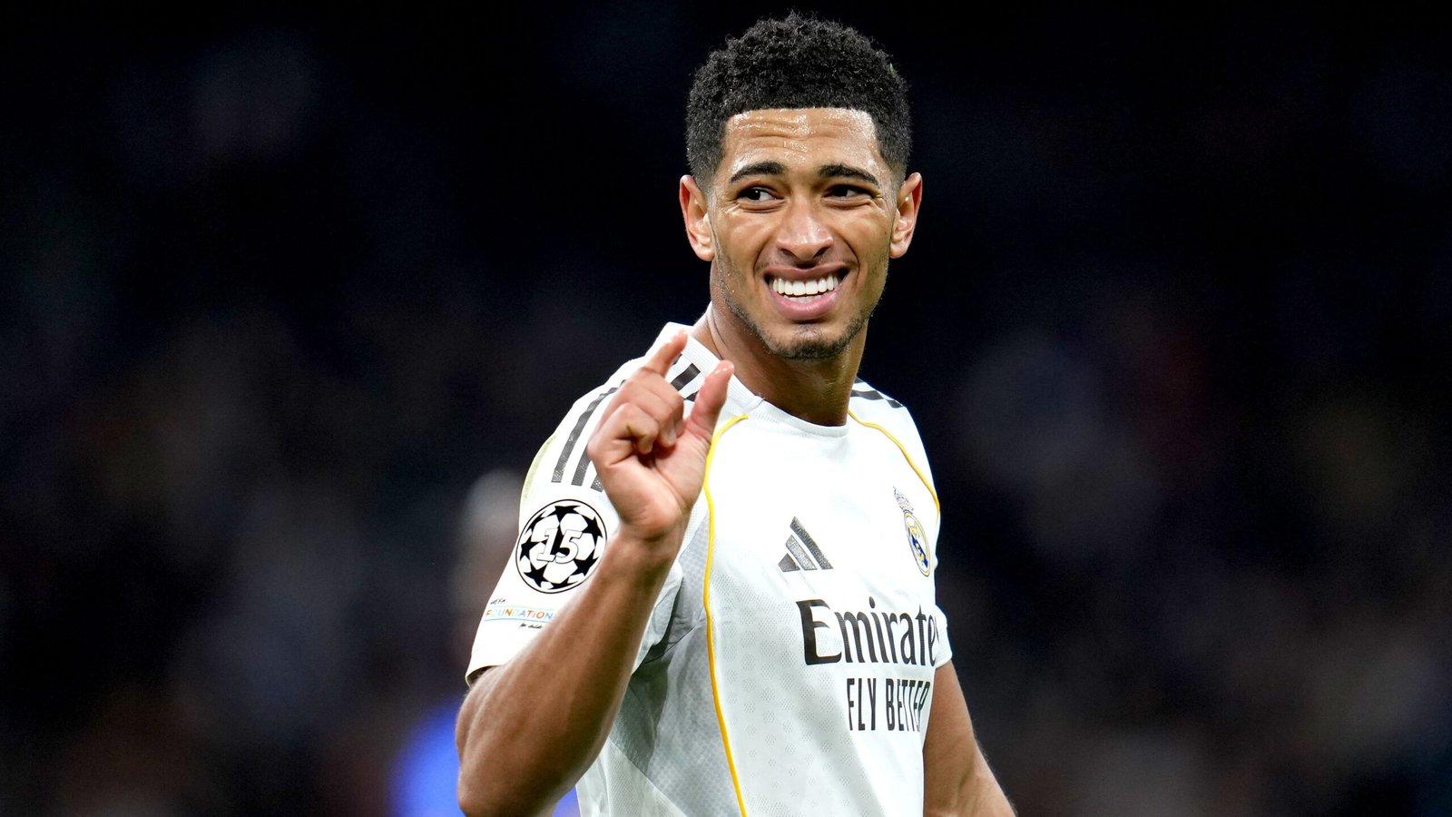 Jude Bellingham, in Real Madrid's white kit, smiles and gestures with his hand during a UEFA Champions League match.