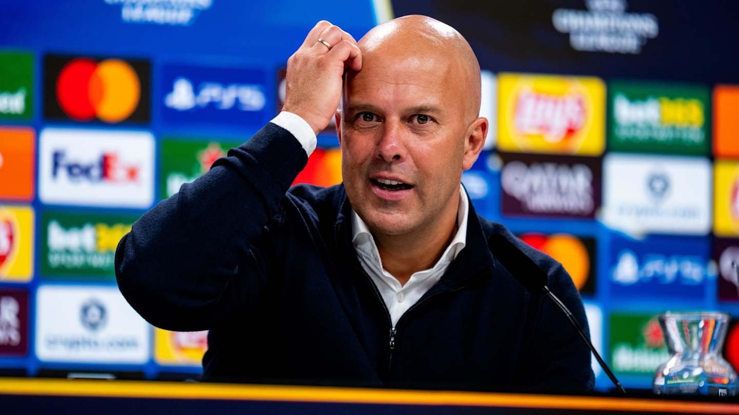 Arne Slot gestures during a press conference, reflecting on Liverpool's transfer plans, with branded logos in the background.