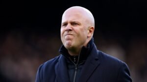 Homme chauve portant un manteau sombre, probablement un entraîneur sportif, avec une expression concentrée.