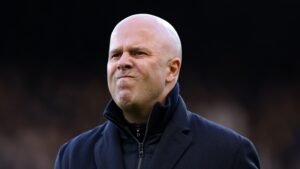 Arne Slot fronsend in een donkere jas, met intense concentratie en bezorgdheid te midden van de wedstrijdactie, wijzend op tactische dilemma's.