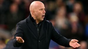 Arne Slot die geanimeerd gebaart vanaf de zijlijn tijdens een voetbalwedstrijd, gekleed in een zwarte jas, met een vervaagde menigte op de achtergrond.