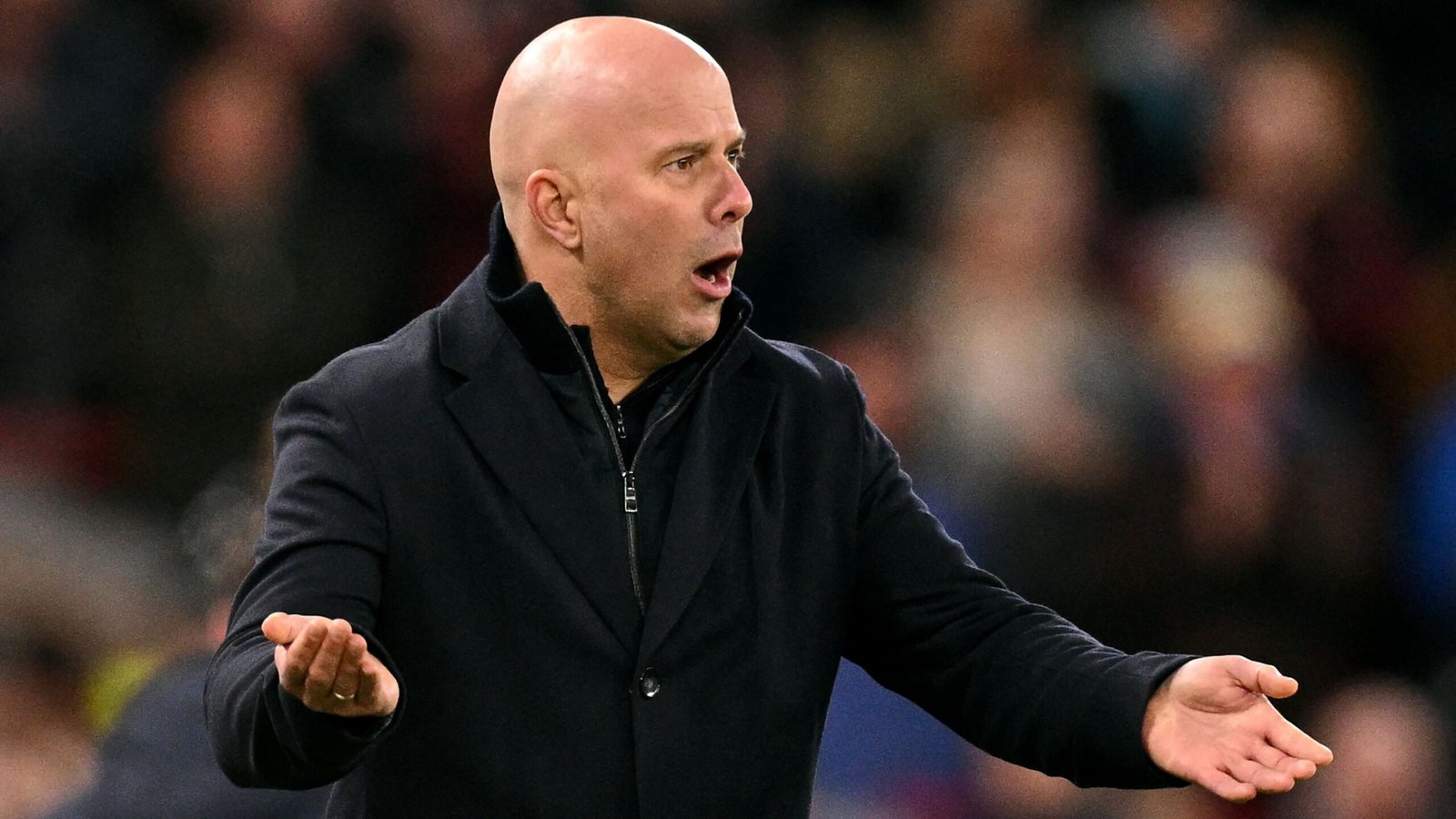 Arne Slot die geanimeerd gebaart vanaf de zijlijn tijdens een voetbalwedstrijd, gekleed in een zwarte jas, met een vervaagde menigte op de achtergrond.