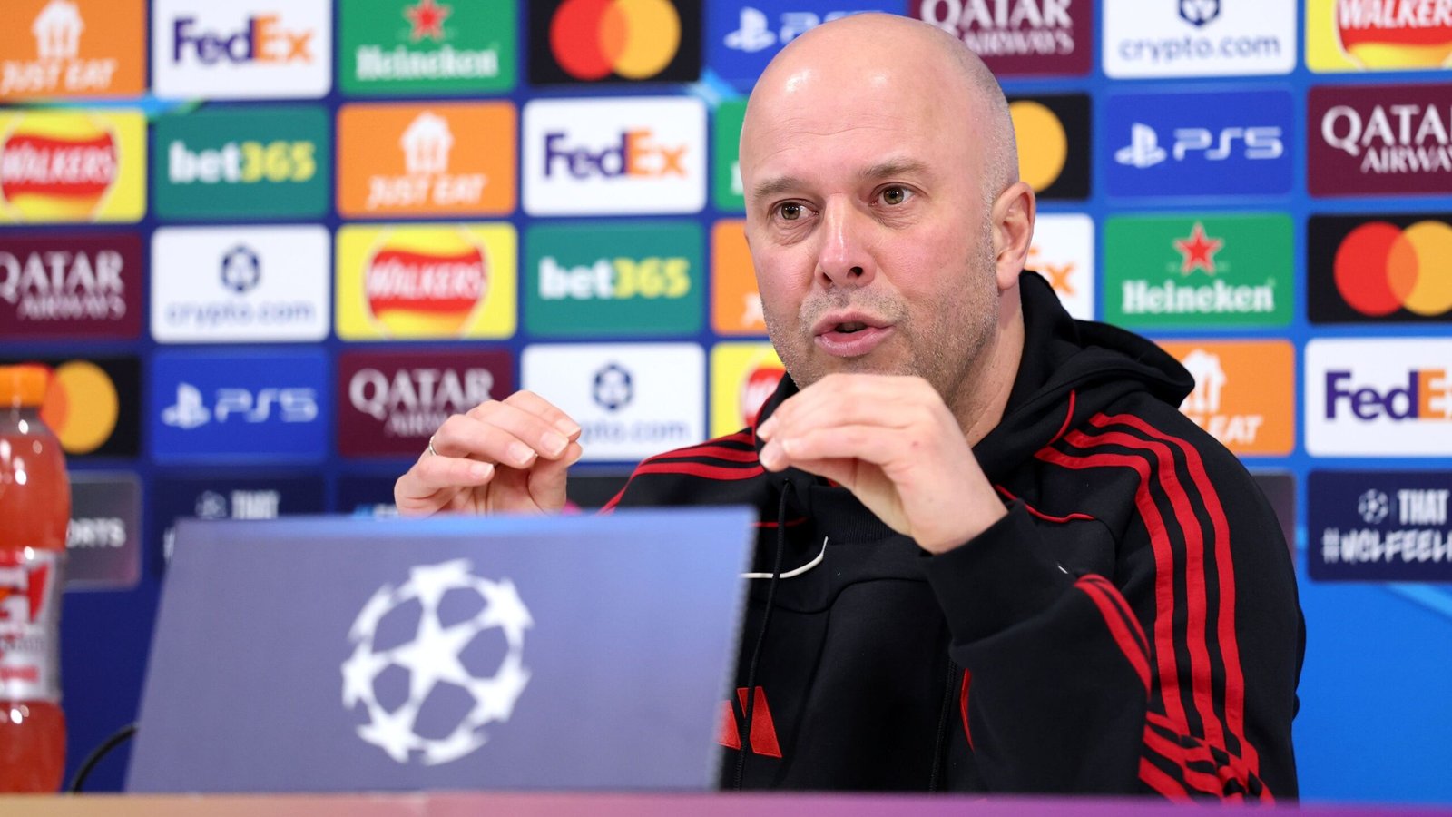 Arne Slot speaks during a press conference, with a UEFA Champions League backdrop featuring various sponsorship logos.