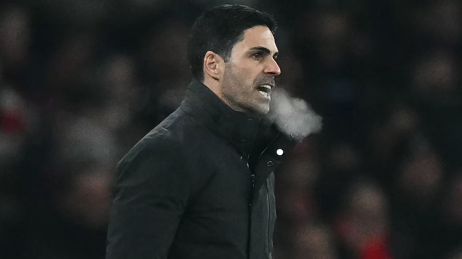 Mikel Arteta, in een donkere jas, instrueert zijn team gepassioneerd vanaf de zijlijn, waarbij zijn adem zichtbaar is in de koude atmosfeer.
