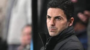 Mikel Arteta kijkt bedachtzaam vanaf de zijlijn tijdens een wedstrijd, gekleed in een zwart jasje tegen een wazig stadionachtergrond.