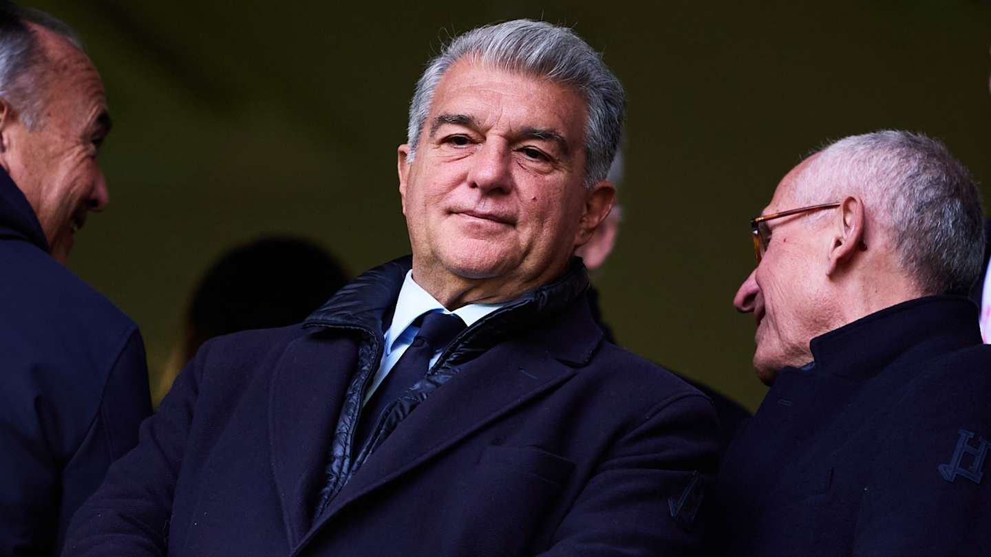 Barcelona president Joan Laporta, dressed in a dark coat, smiles while engaging with two men in suits during a match day.