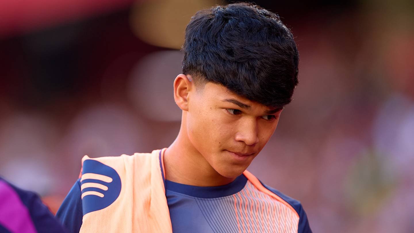 Young football player wearing a navy blue and orange training kit, looking focused as he walks towards the pitch, with a blurred background.
