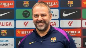 Barcelona manager, smiling in a navy shirt with club logo, backdrop featuring team sponsorships and logos including La Liga and Estrella Damm.