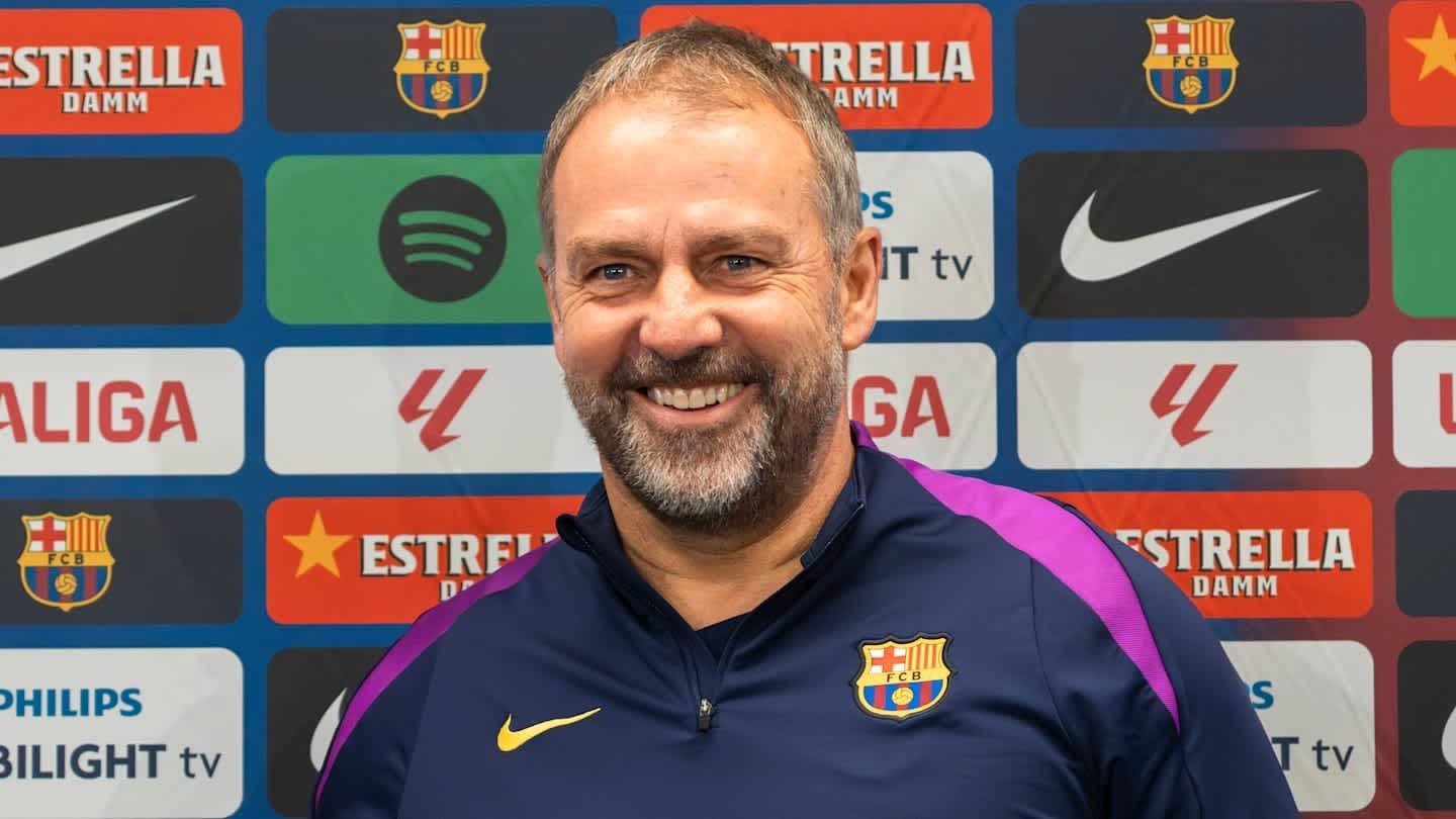 Barcelona manager, smiling in a navy shirt with club logo, backdrop featuring team sponsorships and logos including La Liga and Estrella Damm.