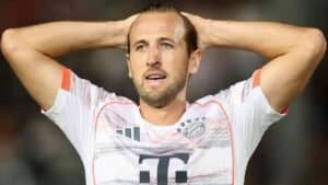 Harry Kane, portant un maillot du Bayern Munich avec des accents orange, semble déçu, se tenant la tête sur le terrain.