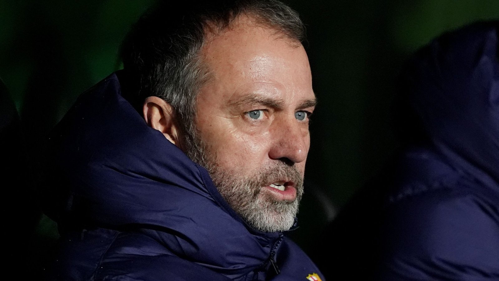 L'entraîneur du Barça affiche une expression intense, portant une doudoune sombre, réfléchissant au match dans un arrière-plan faiblement éclairé.