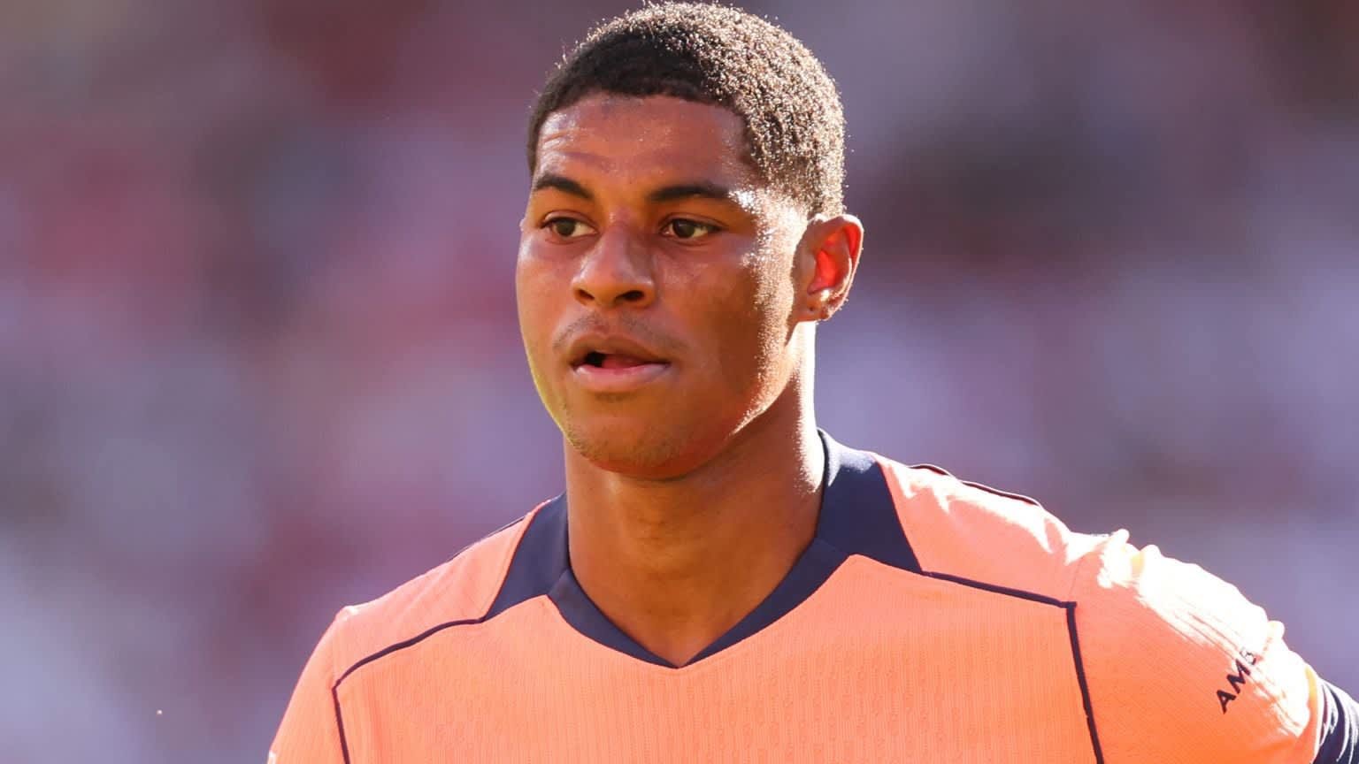 Marcus Rashford portant un maillot de football orange vif, concentré sur le terrain pendant un match, avec un fond de stade flou.