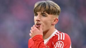 Lennart Karl in het rood van Bayern München, diep in gedachten verzonken tijdens een wedstrijd, met een geconcentreerde uitdrukking en zijn hand aan zijn mond.