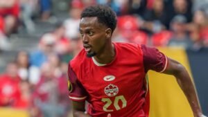 Un footballeur canadien portant un maillot rouge avec le numéro "20" et "CANADA", courant sur le terrain, démontrant de l'intensité devant une foule vibrante.