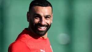 Mohamed Salah sourit pendant un entraînement, vêtu d'un maillot rouge, devant un fond vert flou, incarnant ainsi son esprit joyeux sur le terrain.