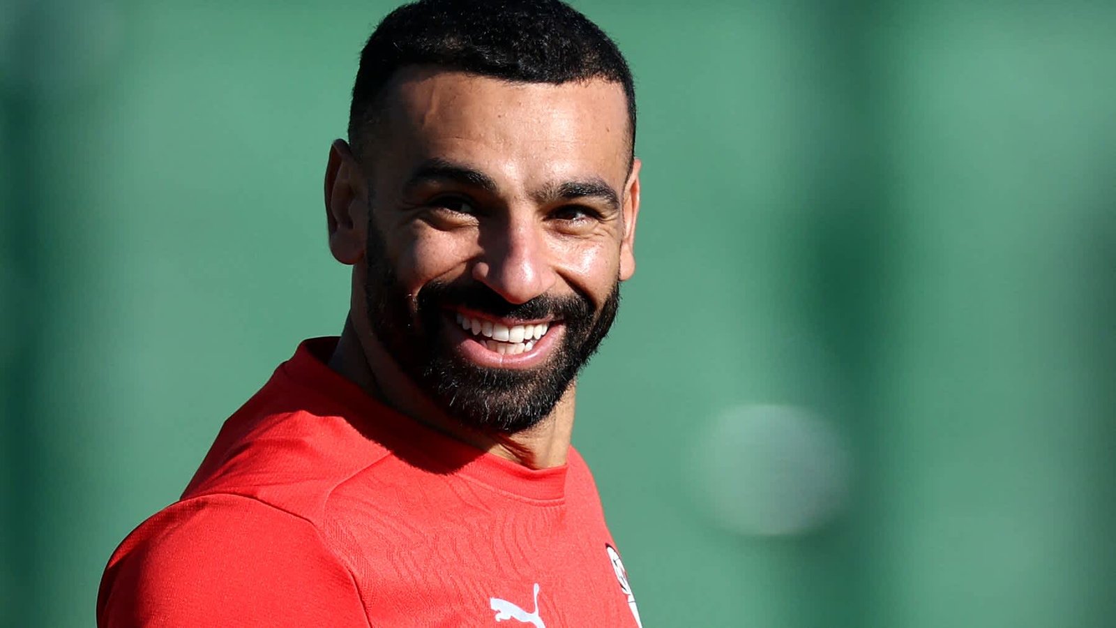 Mohamed Salah sourit pendant un entraînement, vêtu d'un maillot rouge, devant un fond vert flou, incarnant ainsi son esprit joyeux sur le terrain.