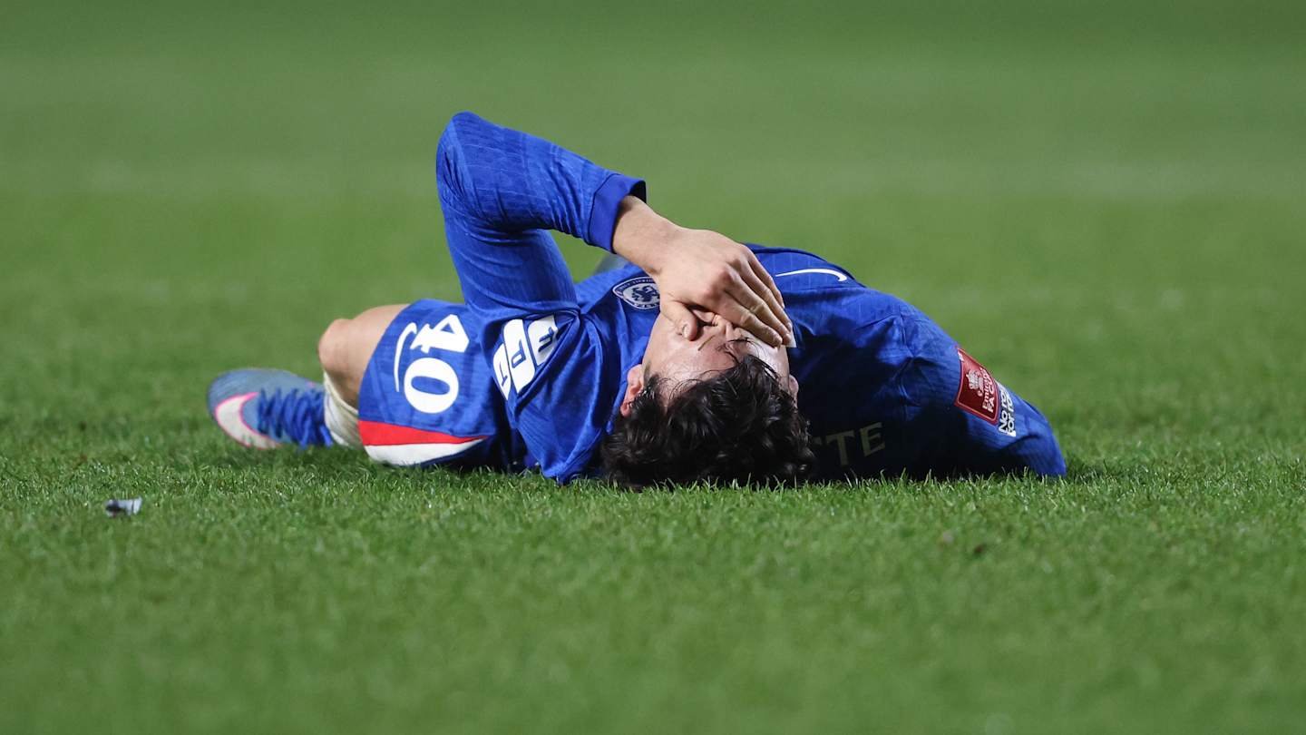 Chelsea player wearing a blue jersey lies on the pitch, covering his face with one hand after a match, displaying signs of distress.