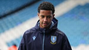 Sheffield Wednesday player in a dark jacket with club crest, walking on a rainy pitch, expressing a focused demeanor amidst a blue backdrop.