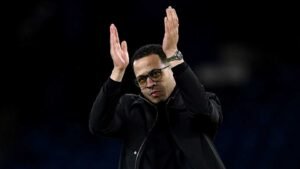 Le manager applaudit les fans les deux mains levées, portant des lunettes et une veste noire, sur fond de stade sombre.