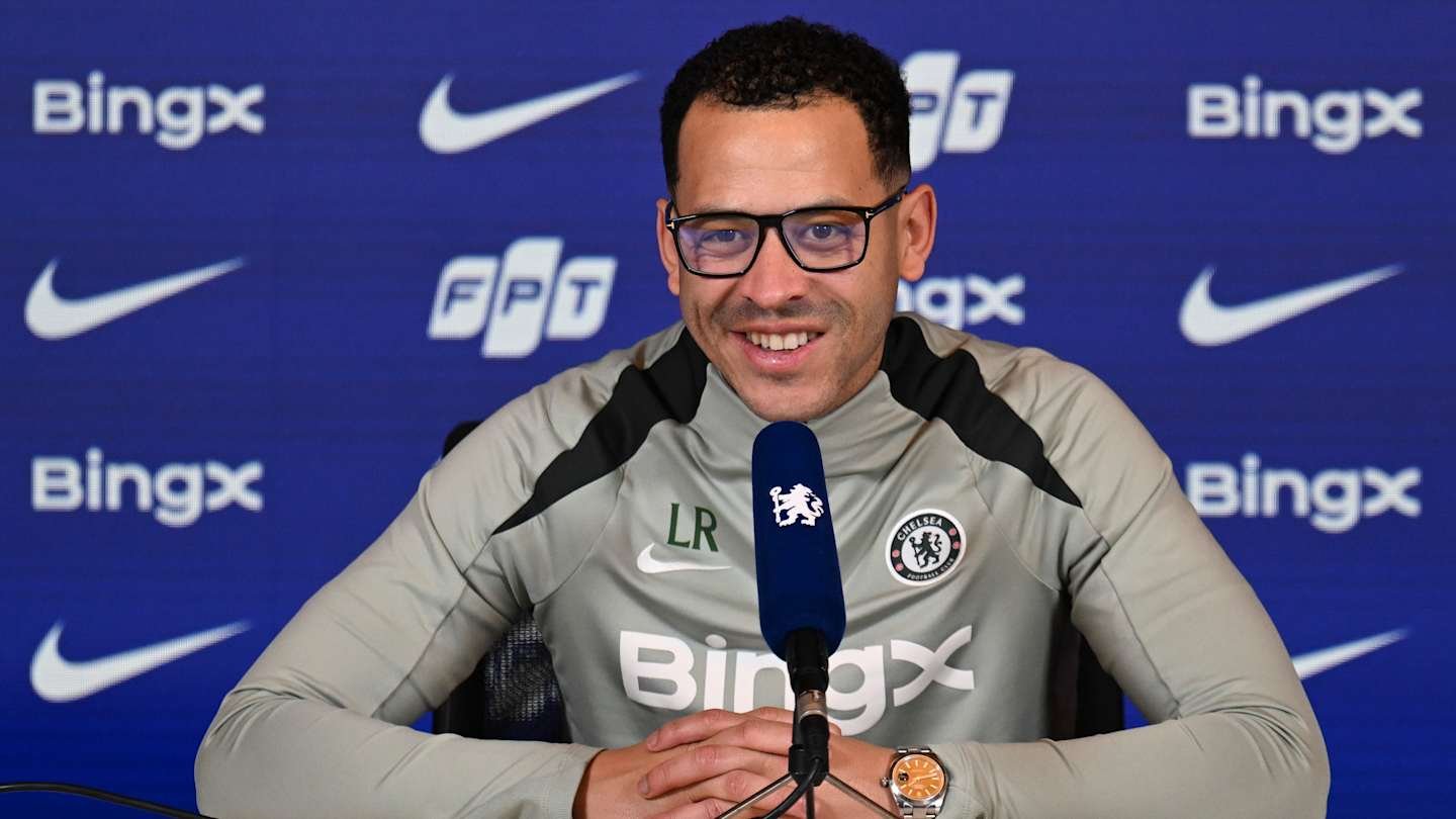 L'entraîneur de Chelsea, vêtu d'un survêtement gris aux logos du club, sourit lors d'une conférence de presse avec un fond arborant les marques Bingx et Nike.