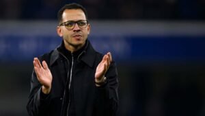 Entraîneur de football marocain applaudissant pendant un match, vêtu d'une veste noire, l'air concentré, avec un stade en arrière-plan.