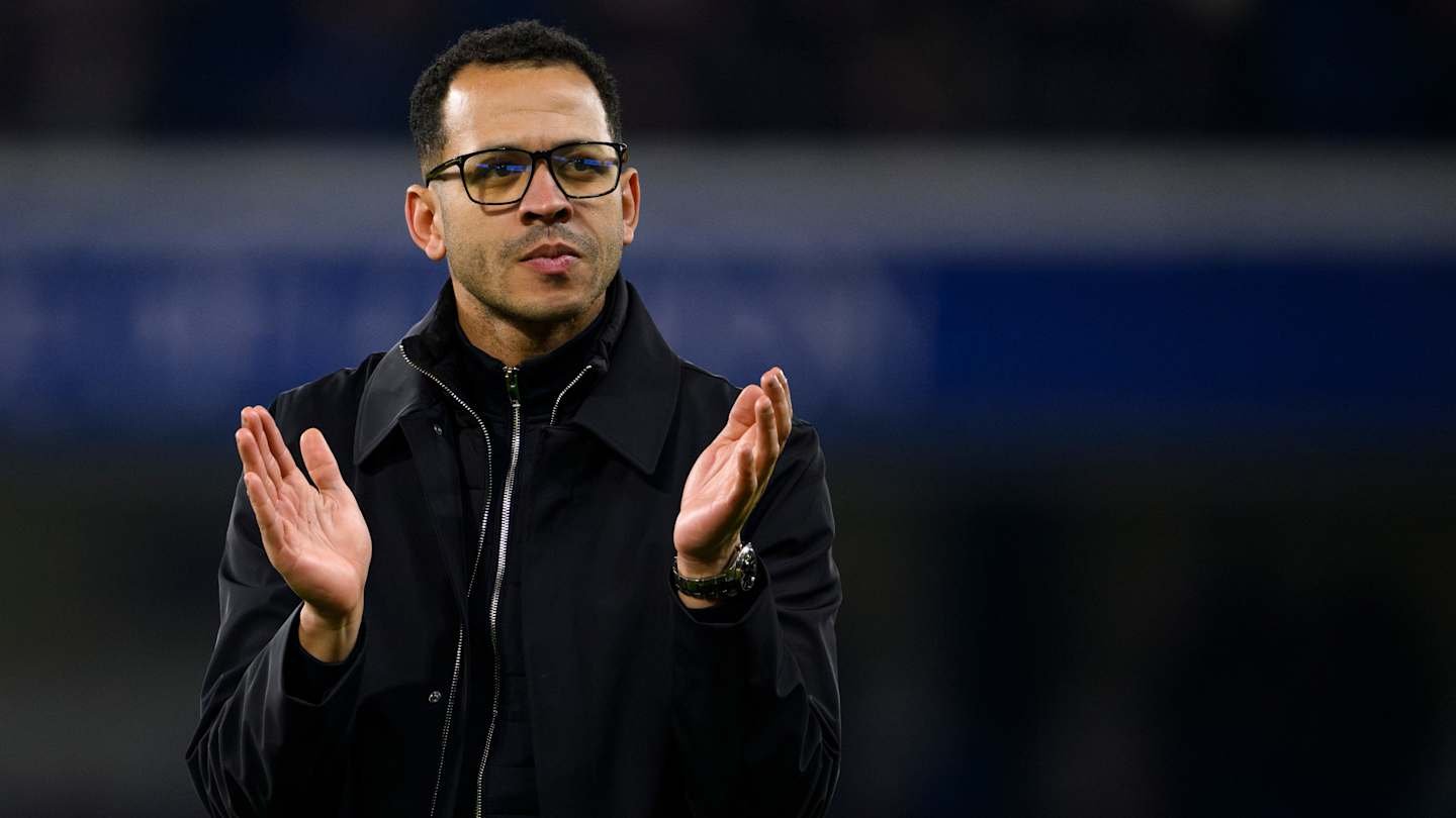 Entraîneur de football marocain applaudissant pendant un match, vêtu d'une veste noire, l'air concentré, avec un stade en arrière-plan.