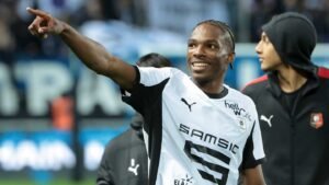 Joueur rennais célébrant avec un sourire, portant un maillot noir et blanc, pointant du doigt les supporters dans le fond de stade.