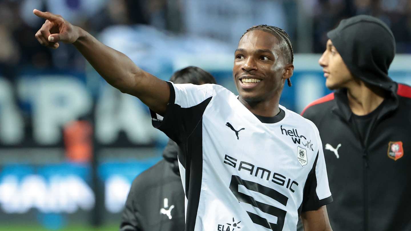 Joueur rennais célébrant avec un sourire, portant un maillot noir et blanc, pointant du doigt les supporters dans le fond de stade.