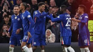 Chelsea players celebrate a goal, wearing blue kits with club logos, as fans cheer in the background during a Champions League match.